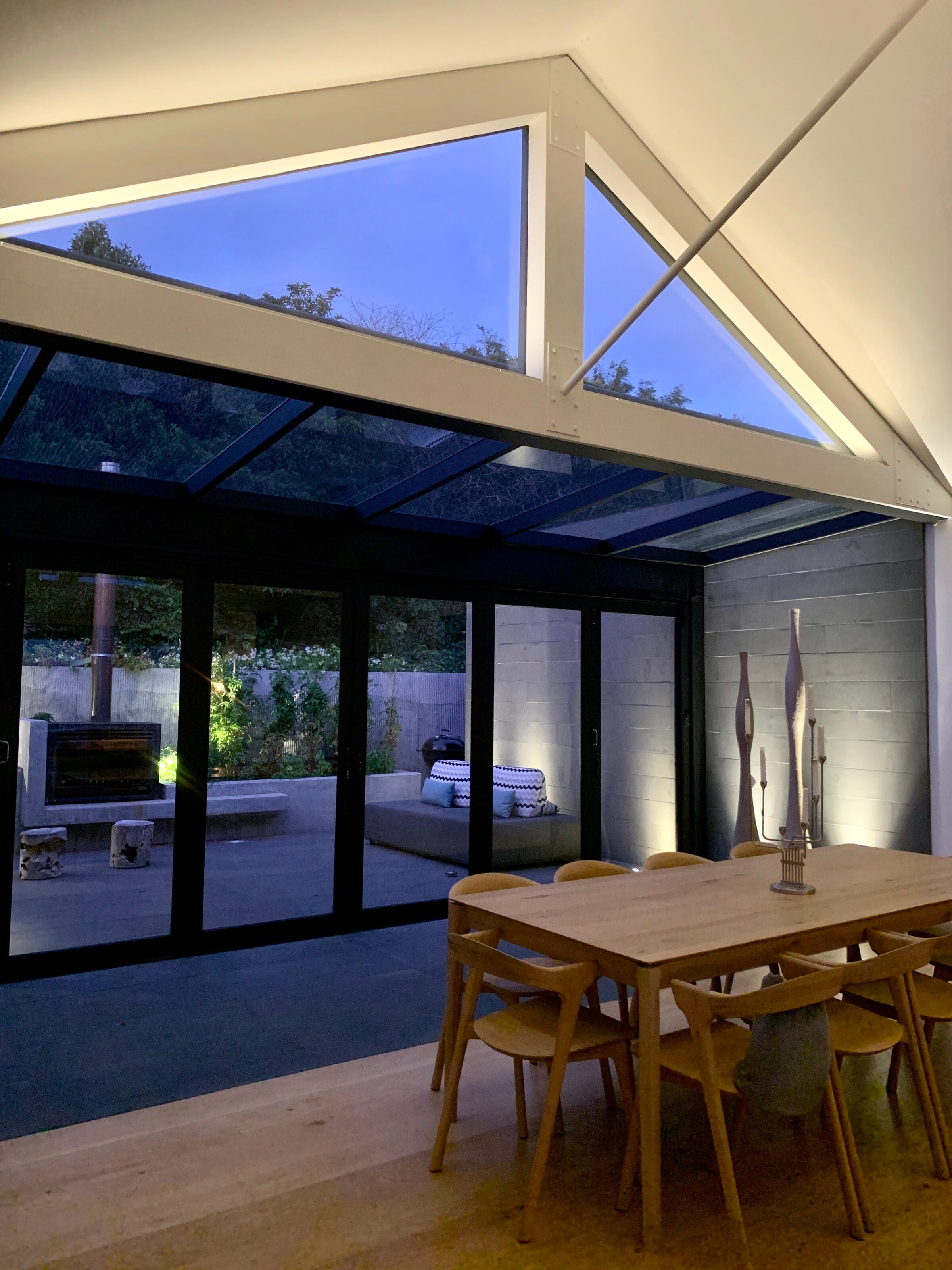 Dining room under a glazed roof at dusk, opening through bifold doors to a courtyard garden, Mount Martha