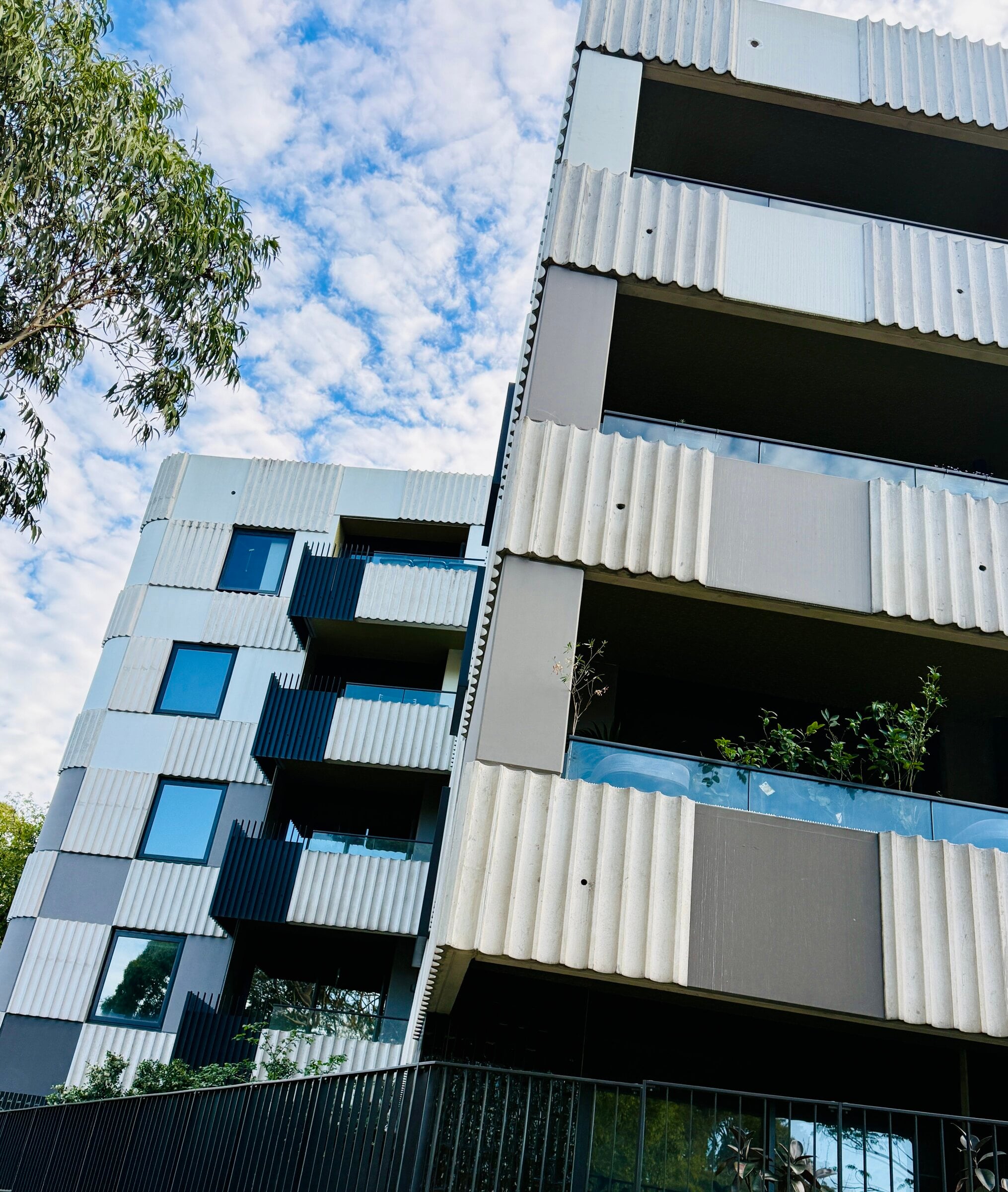 Apartment building with pale ribbed panels and glass balustrades on its balconies, seen from below against a cloudy sky