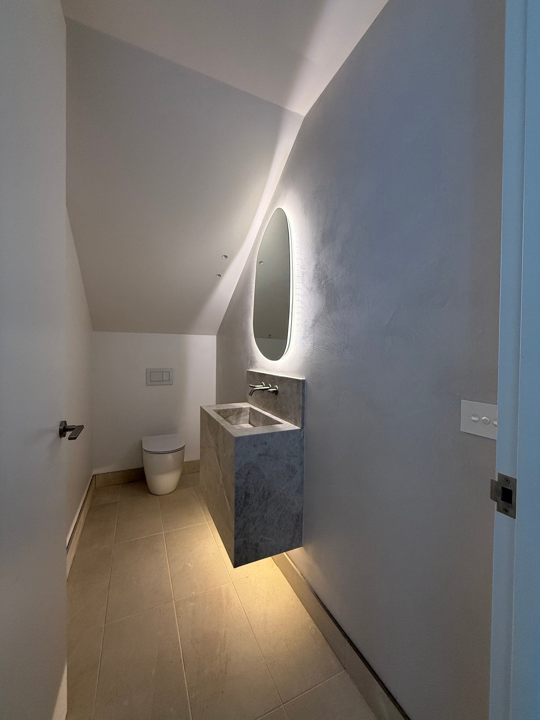 Powder room with a backlit oval mirror over a stone basin under a sloping ceiling, Port Melbourne